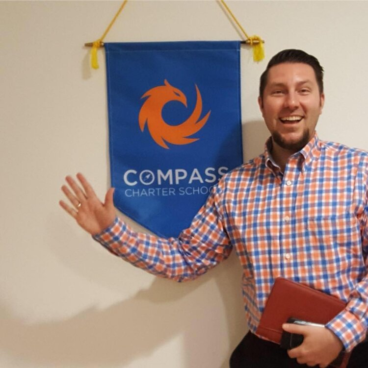 Compass Online Charter School’s Superintendent, J.J. Lewis smiles and waves in front of the Compass flag, eager to support everyone.
