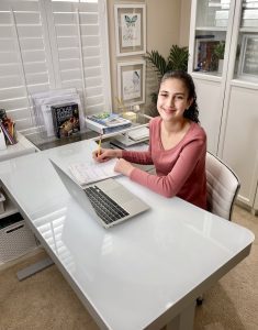A smiling Compass Charter School scholar looks up from her homework in her home office with a laptop open in front of her.
