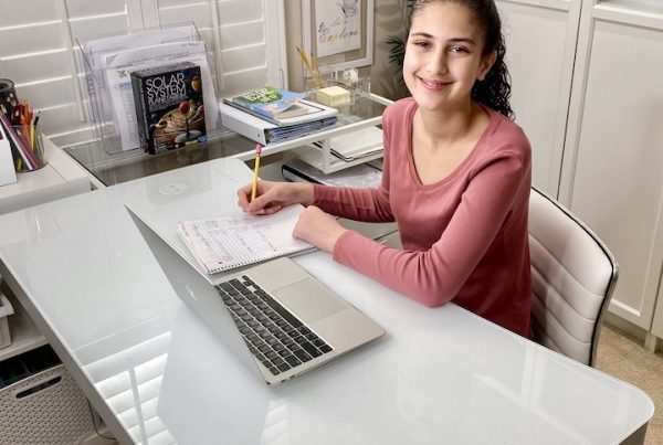 A smiling Compass Charter School scholar looks up from her homework in her home office with a laptop open in front of her.